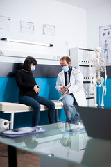 Fototapeta premium In clinic, physician holds digital device to show expectant lady her medical diagnosis during pregnancy checkup. Doctor with tablet sits on hospital bed with pregnant woman, discussing maternal health