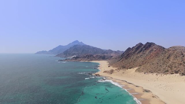 Aerial view of a rugged coastline featuring turquoise ocean waves meeting a sandy beach and rocky mountains on Socotra Island, Yemen.