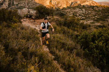Athletic trail runner running along a winding mountain trail surrounded by wild vegetation and rugged terrain. Outdoor endurance sport and adventure lifestyle in nature.