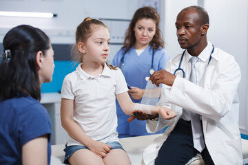 Obraz premium Arm of girl patient being meticulously wrapped with a sterile bandage by multicultural medical personnel. Caucasian youngster accompanied by her mother is being treated by doctor and nurse.