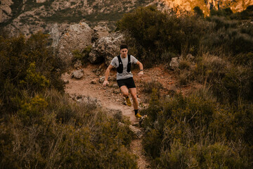 Obraz premium Endurance athlete running along a narrow trail through rugged mountain terrain surrounded by wild vegetation and rocky landscape. Outdoor training session focused on stamina, endurance and performance
