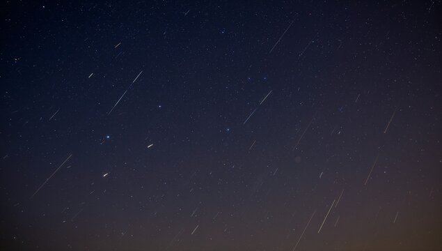 Displaying night sky showing meteor streaks over rural horizon, with stars and faint orange glow