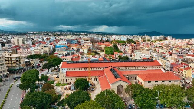 Aerial view of Platia Agoras, a building with a red roof, surrounded by city buildings and lush green trees under a dramatic cloudy sky, Chania, Chania, Greece.