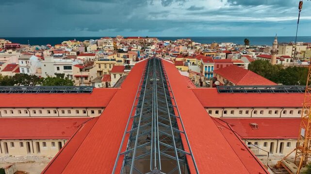 Aerial view of Platia Agoras Market Hall with its striking red roof contrasting against the distant blue sea and sky, Chania, Chania, Greece.