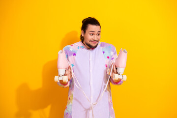 Playful man in pastel pajama jumpsuit holds pink roller skates on bright yellow background