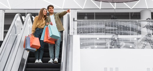 A couple stands on an escalator at a mall, smiling and holding shopping bags. The man points towards something ahead, while the woman looks in his direction. © Prostock-studio