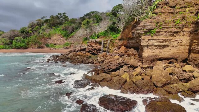 Aerial view of a small, gentle waterfall cascading over rocky cliffs into the ocean, contrasting the rugged rocks with the foamy waves, Tambor, Puntarenas Province, Costa Rica.