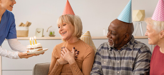 Obraz premium A group of older adults enjoys a birthday celebration in a cozy room. One person serves a cake with candles. Friends smile and share the moment. They wear party hats and show happiness.