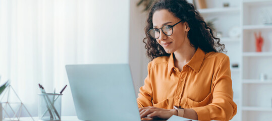 A woman sits at a desk in her home office using a laptop. She looks focused and is dressed in a yellow shirt. Books and plants are visible in the background of the room.