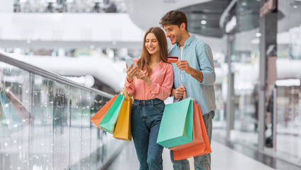 Fototapeta premium Two people are smiling and looking at a phone while walking through a mall. They are holding shopping bags and a credit card. The setting shows a busy shopping area.