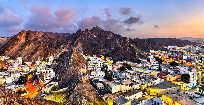 Aerial panorama of the historic Mutrah Fort district in Muscat, Oman, featuring traditional white buildings nestled among rugged mountains during twilight.