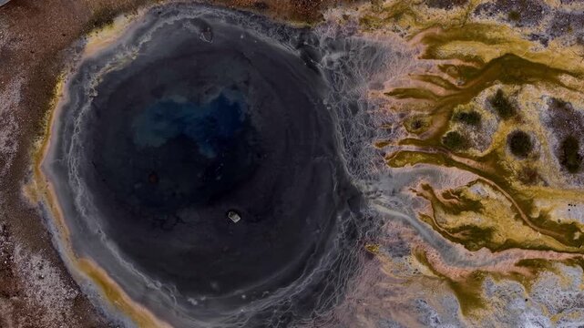 Aerial view of a dark, circular hot spring contrasts sharply with the surrounding colorful, mineral-rich terrain, Hrunamannahreppur, Iceland.
