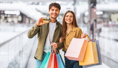 A young couple stands in a mall holding shopping bags and a credit card. They smile while enjoying their shopping experience. The background shows a busy mall with people walking. © Prostock-studio