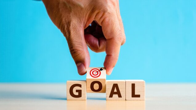 Hand placing wooden cube with target icon on top of 'GOAL' word blocks, symbolizing business objective achievement and success strategy on a blue background