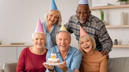 Obraz premium A group of five adults gather in a cozy room to celebrate a birthday. They wear party hats and smile as one person holds a cake with candles. Joy fills the atmosphere.