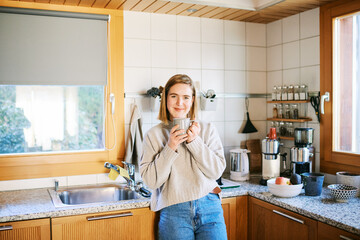 Young woman standing in cozy home kitchen holding warm coffee mug and smiling near window in...