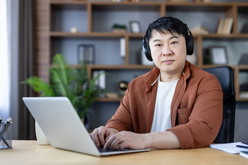 Fototapeta premium Asian man in headphones focused at a home office desk, typing on a laptop while working remotely, attending online meetings and concentrating on productivity and learning