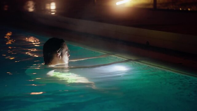 A serene video shows a teenager relaxing in an illuminated swimming pool at night. Underwater lights cast a tranquil green glow, highlighting subtle movements and reflections