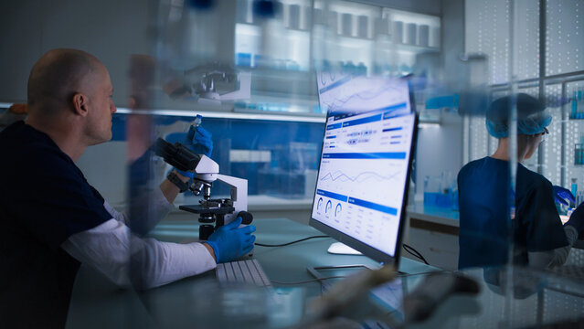 Research work in a lab. Man sitting in front of a computer screen and a microscope testing samples