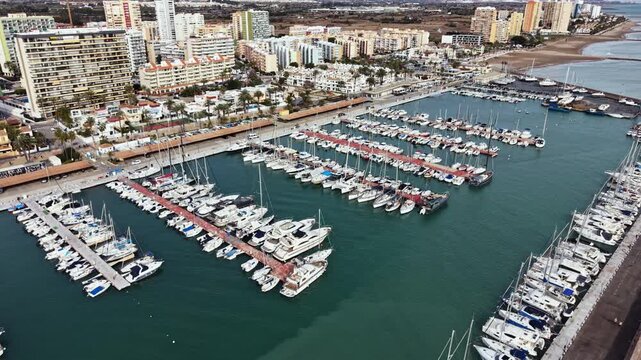 The scene shows a marina with many boats and yachts docked in neat rows. Buildings and a shoreline stretch along the background. Clear skies are above.