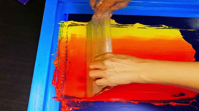 Hands spread warm-toned inks across a screen frame, blending colors from soft yellow to intense red. Close up captures the texture, motion, and artistry involved in crafting vibrant gradient prints.