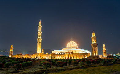 Illuminated view of the Sultan Qaboos Grand Mosque in Muscat, Oman, featuring its golden dome and tall minarets surrounded by landscaped gardens at night.