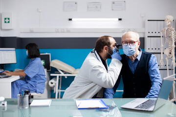 Naklejka premium Young doctor uses otoscope on elderly sick man seated in clinic, ensuring accurate diagnosis during health consultation. Male otologist doing ear examination on pensioner patient with medical tool.