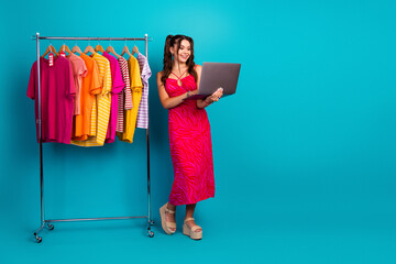 Young chic female model stands beside a colorful clothing rack while holding a laptop in a bright...