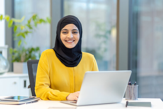 Muslim businesswoman wearing a black hijab and yellow blouse, smiling while working on a laptop at her desk in a modern office, representing professionalism and diversity in the workplace