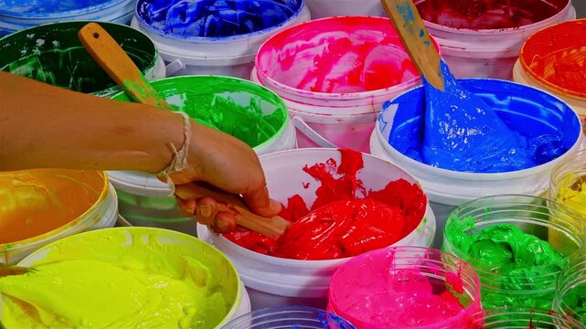 On a wooden workbench in a screen printing shop, several plastisol ink cans are open, revealing rich tones soft pastels, strong primaries, and deep shades. A wooden spoon and trowel rest nearby.
