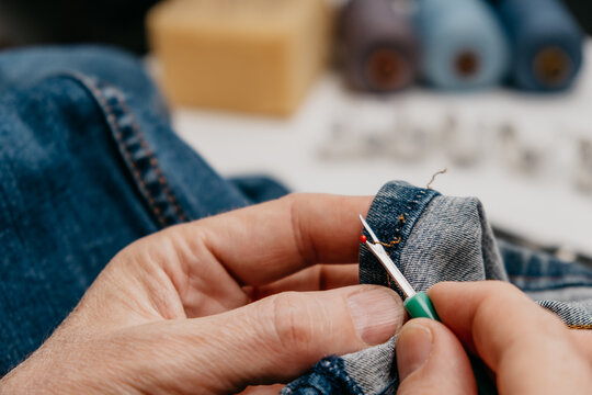 tailor is using green seam ripper to take apart stitches on denim fabric. scissors and sewing supplies are present on table. setting shows crafting area with tools. closeup.