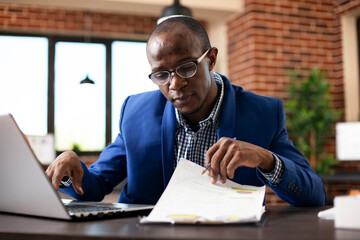 Black male analyst reading company research information on clipboard and typing on his laptop....