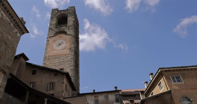 Campanone Torre Civica bell tower with clock in Bergamo Italy, static low angle shot over old rooftops as bright summer daylight lights rough stone walls on historic Piazza Vecchia square