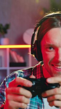 Leisure concept where man in headphones pressing buttons on joystick when playing video racing and raising his hands after victory on the background of home interior in the evening