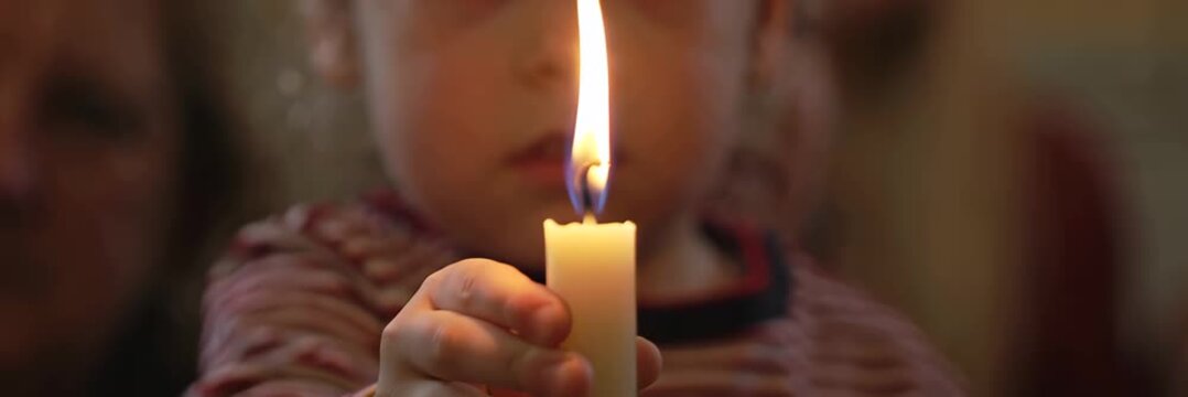 Child holding burning candle close to face in dark environment, quiet emotional moment and spiritual reflection concept in wide banner format for website.