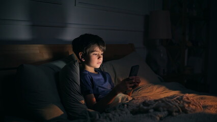 Fototapeta premium Little kid looking cellphone lying dark bedroom closeup. Boy staring smartphone