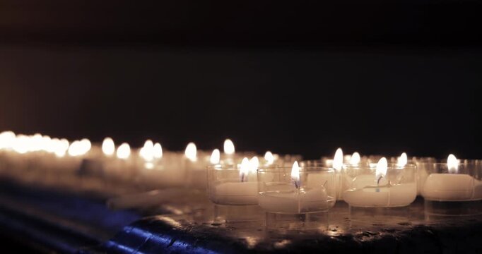 Close focused prayer candles on polished marble ledge, static view holds on bright wicks and wax pools in the foreground as distant flames dissolve into creamy bokeh inside a shadowy sanctuary
