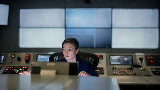 An operator in soft normal lighting views multiple screens on a control wall with focus on facial expression while control panels in the foreground remain out of focus.