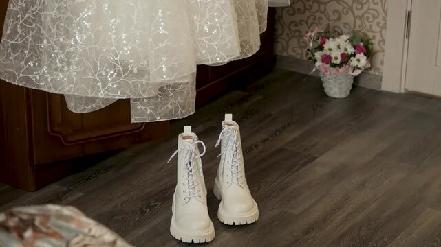 Modern white leather platform combat boots standing on dark laminate floor beneath hem of sparkling lace wedding dress with basket of flowers in background