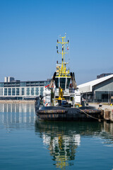 Fototapeta premium a black, white, and yellow tugboat moored at a concrete pier, reflecting perfectly in the calm harbor water, with modern waterfront buildings in the background