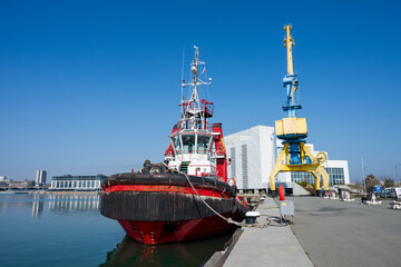 Fototapeta premium a red and black tugboat moored at a concrete dock next to a massive yellow and blue portal crane, with a modern white building and the city skyline