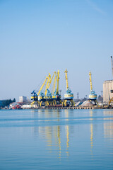 Obraz premium a busy industrial port featuring a row of yellow gantry cranes and a concrete grain elevator reflecting in the calm harbor water under a clear blue sky.