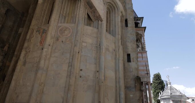Low angle glide from Santa Maria Maggiore basilica wall to Bergamo Baptistery in Piazza Duomo, the camera pans along pale stone buttresses and faded fresco circles