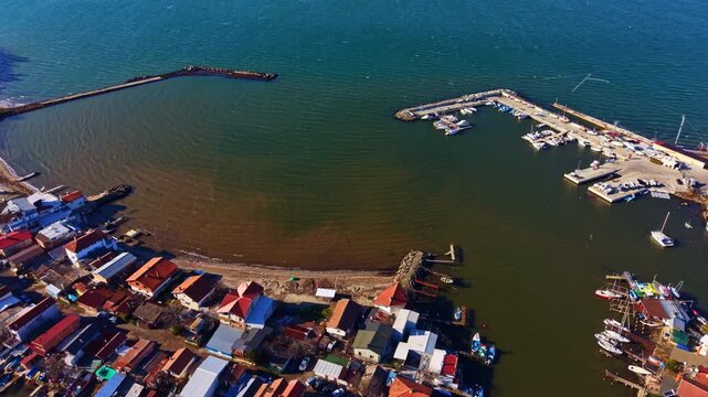 A view of a harbor with boats docked next to small buildings. The water is calm, and the sun is shining. Several vessels are lined up at the shore.