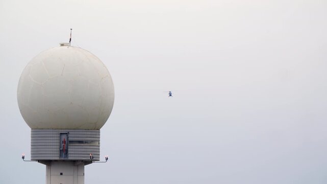 A helicopter flies through a hazy sky, passing behind a large white radome of an air traffic control tower at an airport. Static long shot of aviation technology and surveillance