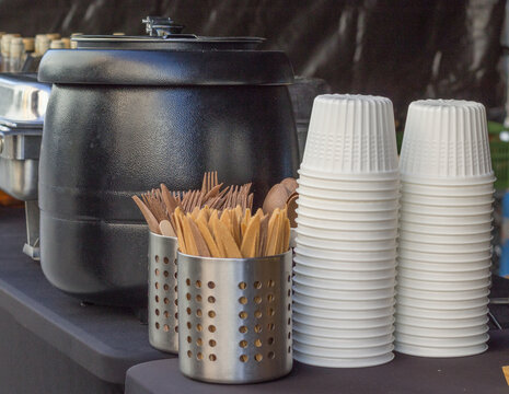 Stoisko z gorącym napojem na wynos, termosy i stos kubk&oacute;w jednorazowych, drewniane mieszadełka, catering w plenerze.