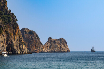Naklejka premium Scenic view of Cilvarda Burnu cape and steep rocky cliffs seen from Cleopatra Beach. Mediterranean coastal landscape with tourist boats sailing near sea caves in Alanya, Turkey.