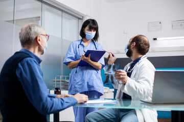 Obraz premium Old man sitting in clinic and listening to young healthcare workers discuss his lung radiograph and treatment options. Doctor speaking to nurse with clipboard as senior patient awaits xray explanation