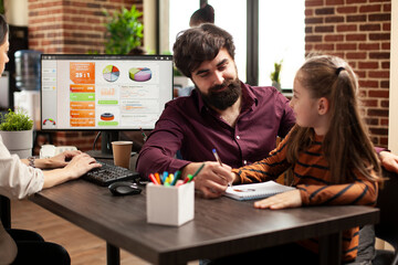 Obraz premium Bearded man drawing on notebook with daughter of coworker at desk in startup company. Working mother using computer as male colleague plays with her little girl during office hours in brick wall room.
