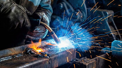 Close-up of a skilled welder's hands in protective gloves operating a welding torch, creating bright blue sparks and molten metal in an industrial workshop.